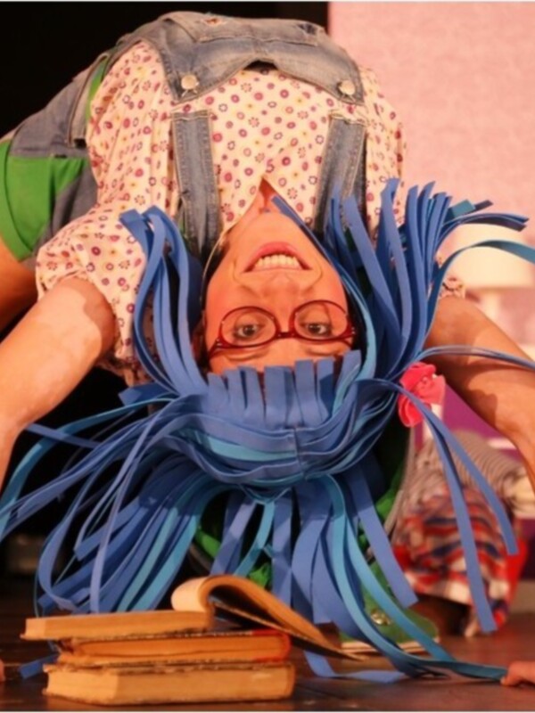 Una figura de papel con cabello azul y gafas rojas, levantando un par de zapatos en una escena teatral.