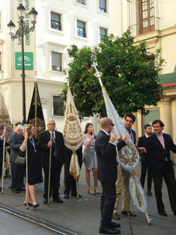 Una procesión con personas vestidas formalmente, ondeando banderas y sosteniendo estandartes en una calle con edificios históricos y árboles verdes.