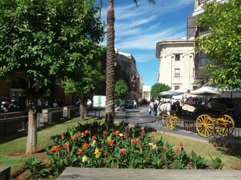 Imaginación: Un hermoso parque con flores coloridas, árboles verdes y un palo de madera en el suelo. El cielo es azul con algunas nubes blancas. La imagen parece ser una ilustración o una representación artística de un parque natural.