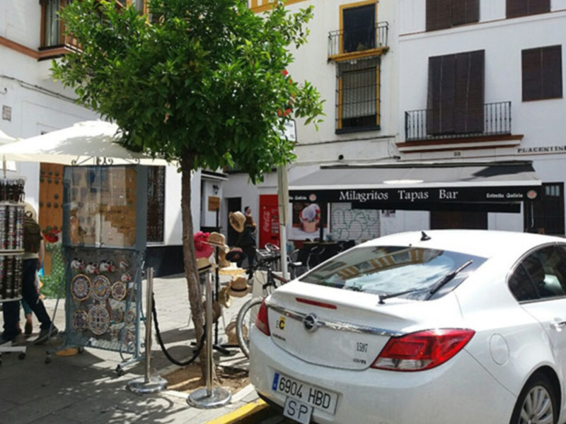 Callejón urbano con árboles, tiendas, y un bar llamado 'Milagritos Tapas Bar' en un edificio blanco con balcones de hierro.