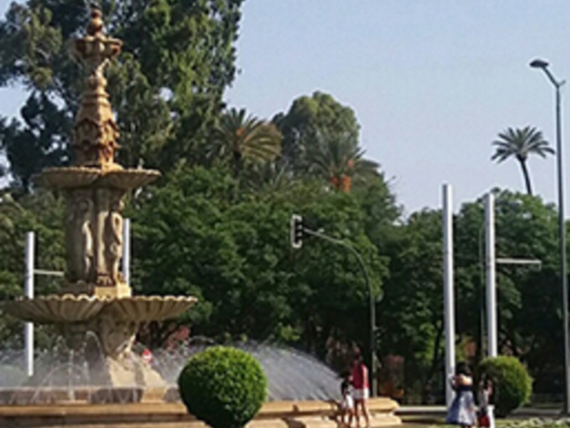 Fuente ornamental en un parque con árboles y personas caminando.