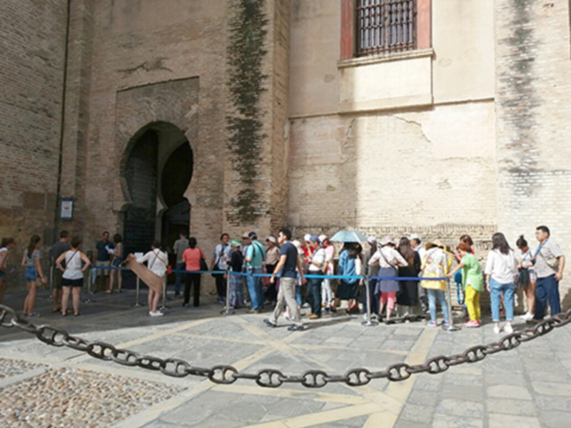 Una fila de personas esperando en una plaza con una estructura histórica en el fondo, rodeada de una cerca metálica.