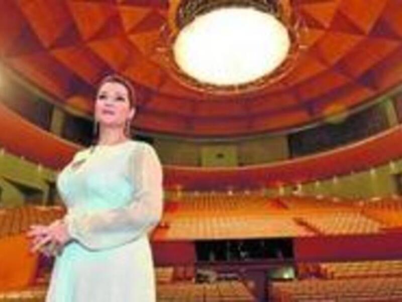 Una mujer en un vestido blanco se encuentra en el centro de una sala de conciertos con techo dorado y asientos naranjas. La iluminación es cálida, creando un ambiente acogedor y profesional para una presentación o concierto.