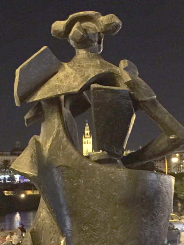 Escultura de bronce en la noche, con un fondo nocturno que incluye una torre iluminada y árboles verdes. La escultura parece ser una figura femenina con un sombrero y una prenda de vestir. El ambiente es谧谧，暗示着一种宁静和优雅。