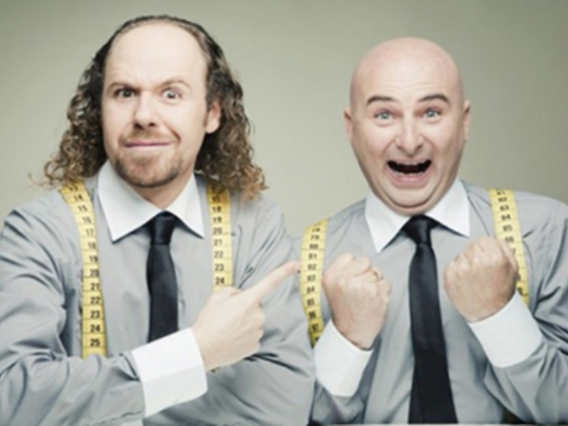 Dos hombres en trajes con cintas métricas, expresando emoción y celebración.