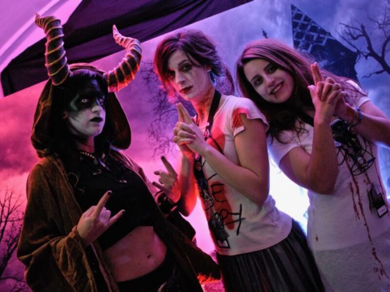Tres personas disfrazadas en un evento temático, posando con gestos de rock y una atmósfera de Halloween en el fondo.