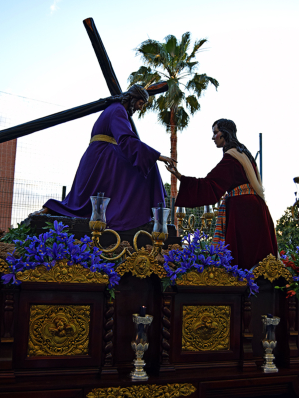 Imaginaria escena de procesión religiosa con estatua de Jesús en la cruz y figura vestida de rojo, rodeados por flores azules y palmeras.