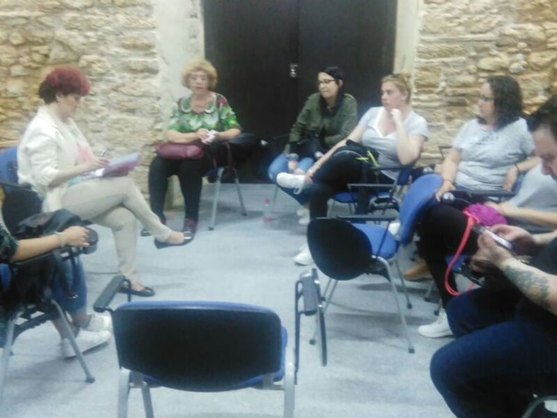Una reunión de trabajo en una sala con paredes de piedra, personas sentadas en sillas, y una persona en el centro escribiendo en una hoja de papel.