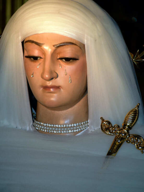 Una imagen de la Virgen María con una expresión triste, rodeada de una capa blanca y una corona dorada. Las lágrimas son visibles en su rostro, creando un sentimiento de dolor y compasión.