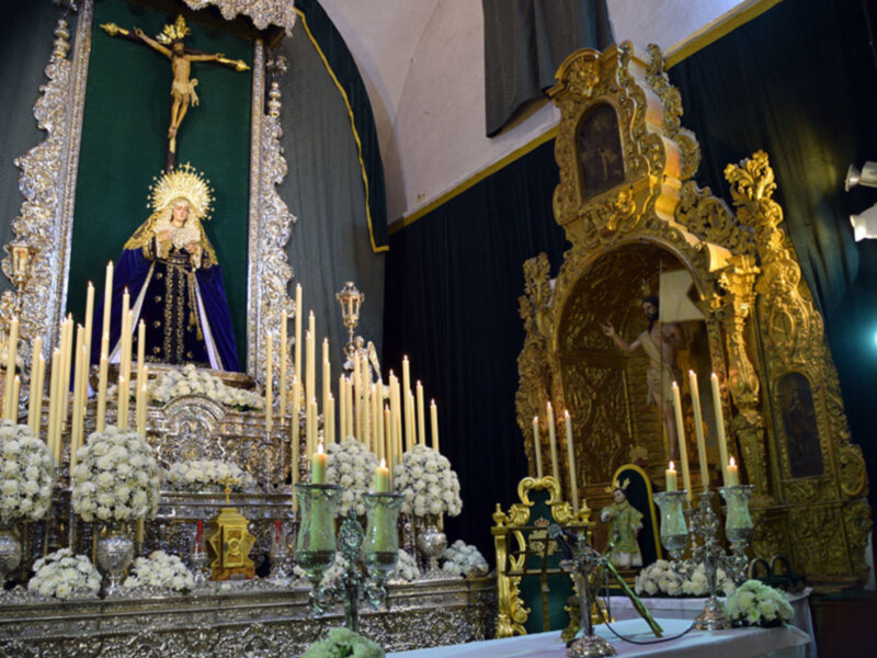 Imagen de un altar con una imagen de la Virgen María y el Cristo crucificado en el centro. Alrededor, hay velas encendidas y flores blancas. El altar está rodeado por adornos dorados y una araña de cristal en el frente. La imagen se ilumina con luces adicionales, creando un ambiente de reverencia y devoción.