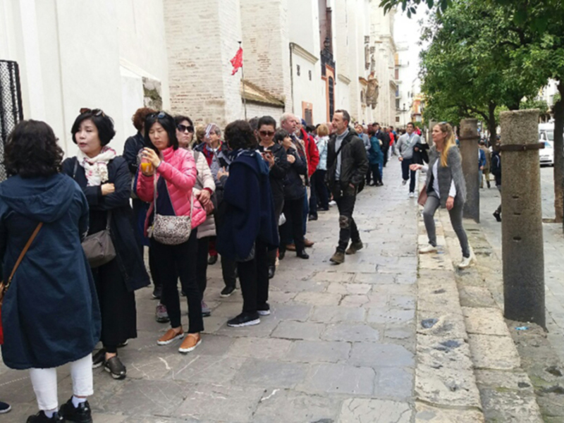 Una multitud de personas camina por una calle adoquinada, con edificios blancos y rojos al fondo. Algunas están hablando, mientras otras observan el entorno. La imagen muestra un día soleado con cielo claro y algunas hojas verdes en el lado derecho.
