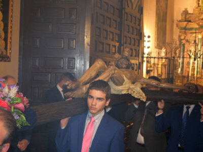 Procesión religiosa con la imagen del Cristo de la Buena Muerte en una iglesia, acompañada por fieles en trajes formales.