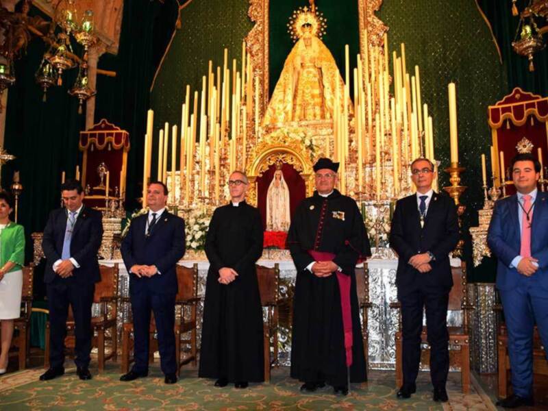 Una imagen de una ceremonia religiosa en un templo con una estatua dorada de la Virgen María y numerosas velas encendidas. En primer plano, seis hombres vestidos formalmente observan la estatua, algunos con corbatas y otros con chalecos. La decoración incluye cruces y mantos dorados, creando un ambiente solemnemente reverente.