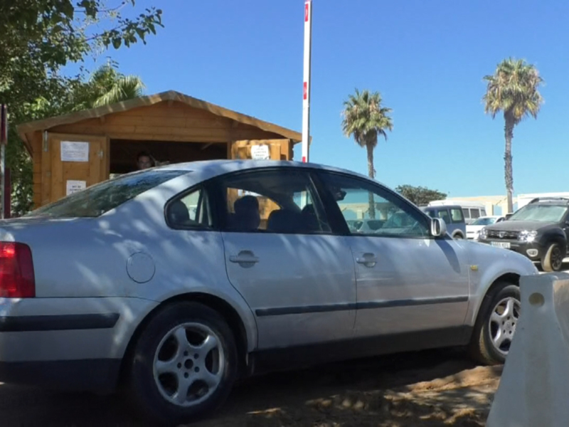 Un coche blanco estacionado en un aparcamiento con una caseta de madera y palmeras al fondo.