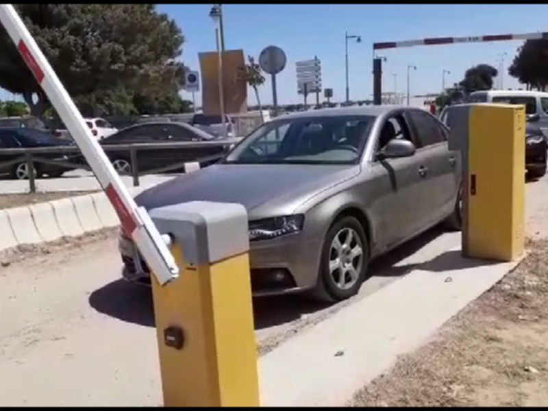 Un coche gris está estacionado en una plazuela amarilla, con un sistema de control de acceso en el fondo. La imagen muestra una vía pública con otros vehículos y edificios alrededor.