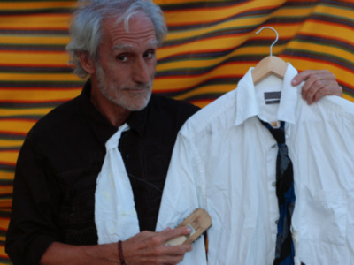 Un hombre con cabello gris y barba está sosteniendo una camisa blanca sobre un perchero, frente a un fondo de tela con rayas naranjas y amarillas.