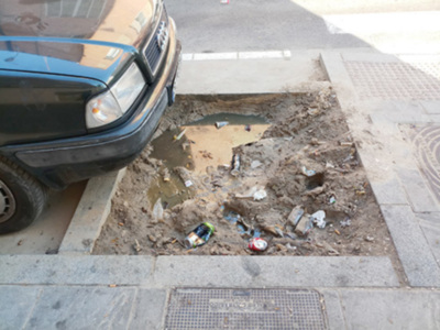 Se observa un coche estacionado en una calle con desperdicios y basura acumulados en el suelo, cerca de un agujero en la calzada. La escena sugiere problemas de limpieza y mantenimiento en el área.