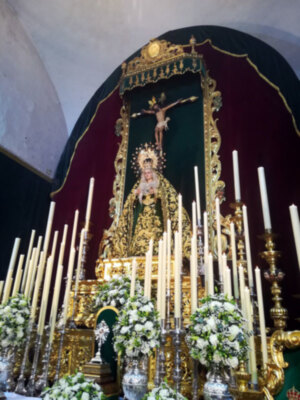 Altar con imagen de la Virgen y Cristo crucificado, adornado con flores y candelabros, en un entorno religioso.