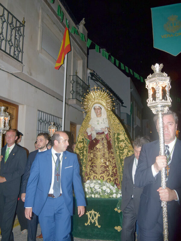Imagen de una procesión religiosa nocturna con una imagen venerada en medio, rodeada por hombres en trajes formales y banderas.
