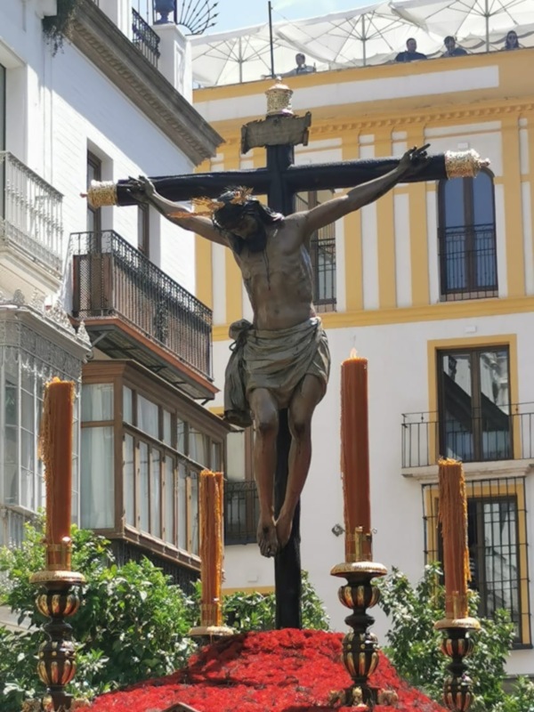 Imagen de la Semana Santa en España, con un Cristo crucificado en una procesión. Fondo de edificios históricos y flores rojas al pie del Cristo.