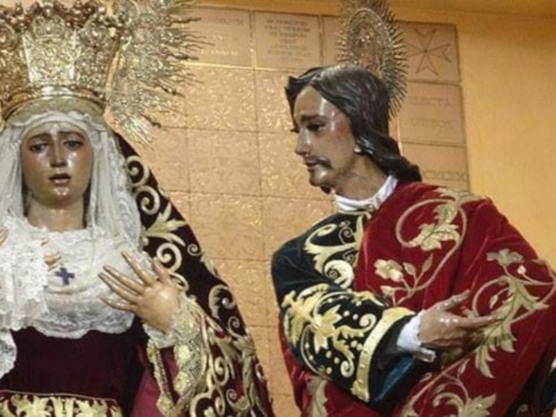 Dos estatuas religiosas, una vestida de blanco con un manto y tocado con un halo, y la otra con traje rojo y dorado. Ambas están en una iglesia o templo, con un muro de fondo que tiene inscripciones.