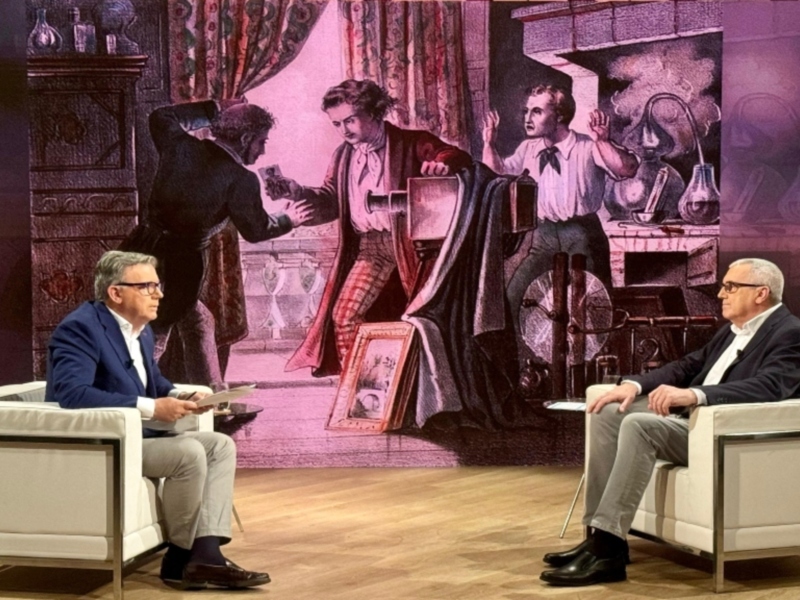 Dos hombres sentados en sillones blancos en una sala con un fondo de ilustraciones antiguas, uno de ellos sostiene un libro abierto. El ambiente es formal y sofisticado, con detalles de estilo clásico en el fondo.