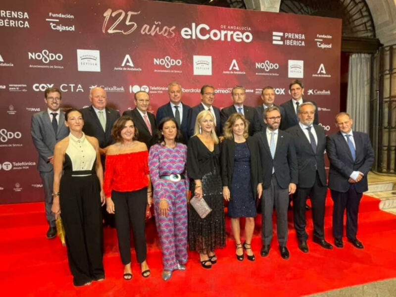 Una imagen de una celebración formal con un grupo de personas posando en un escenario rojo. La imagen muestra a una mujer vestida con un vestido rojo, otra con un vestido azul y estampado, y otros individuos en trajes. La imagen está firmada con el texto "125 años" y "elCorreo", junto con logos de empresas.