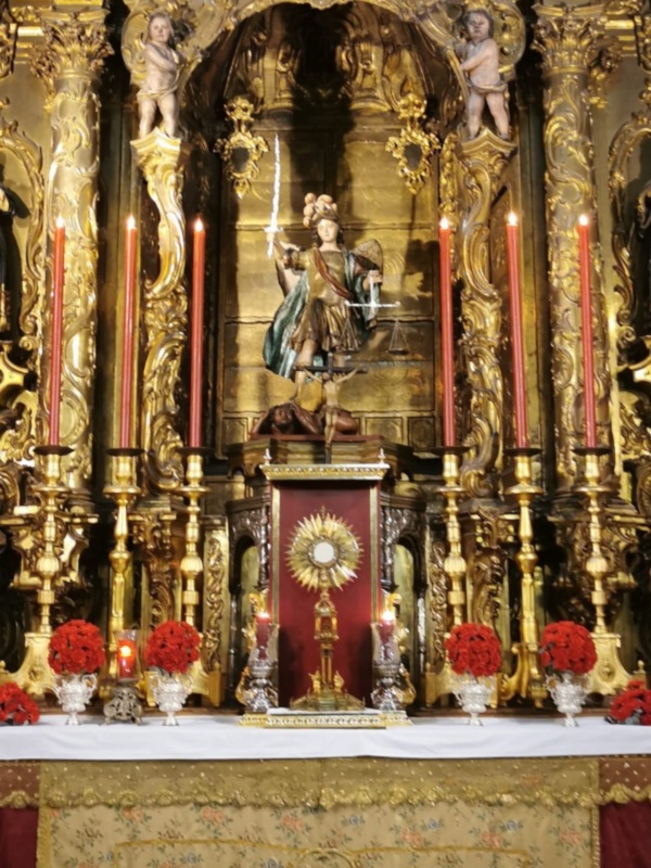 Altar barroco con estatuas, candelabros y flores rojas, decorado con oro y detalles escultóricos.