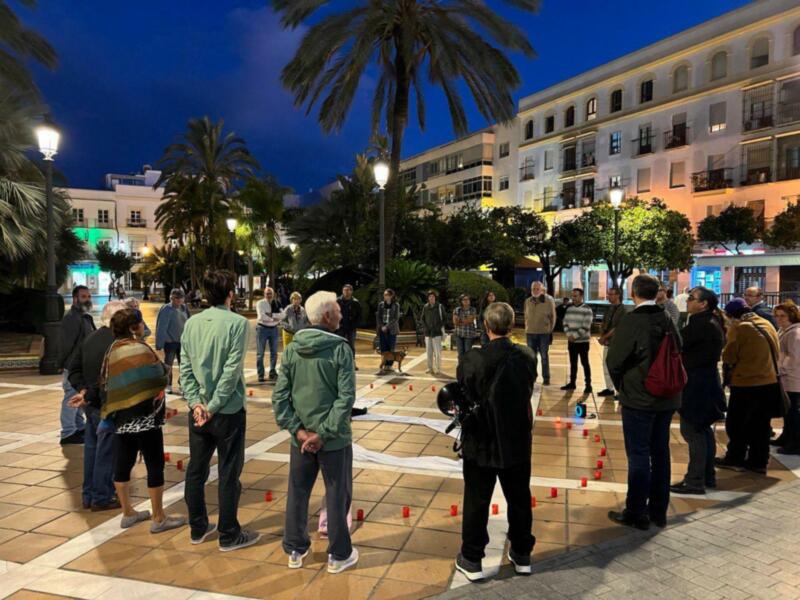 Un grupo de personas se reúne en un espacio público iluminado por farolas, rodeado de palmeras y edificios con ventanas iluminadas. La noche cae, creando un ambiente tranquilo y acogedor.
