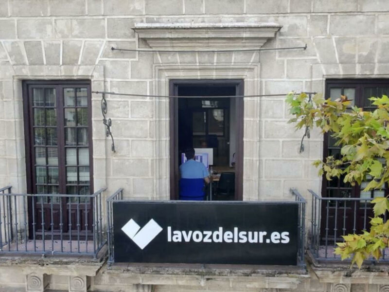 La entrada de un edificio con una puerta principal y dos ventanas laterales. En el centro, hay una persona dentro de la casa, y en la parte inferior se puede ver un letrero con el nombre "lavozdelsur.es". El edificio tiene una fachada de ladrillo y una pequeña ventana con barrotes en la parte superior.