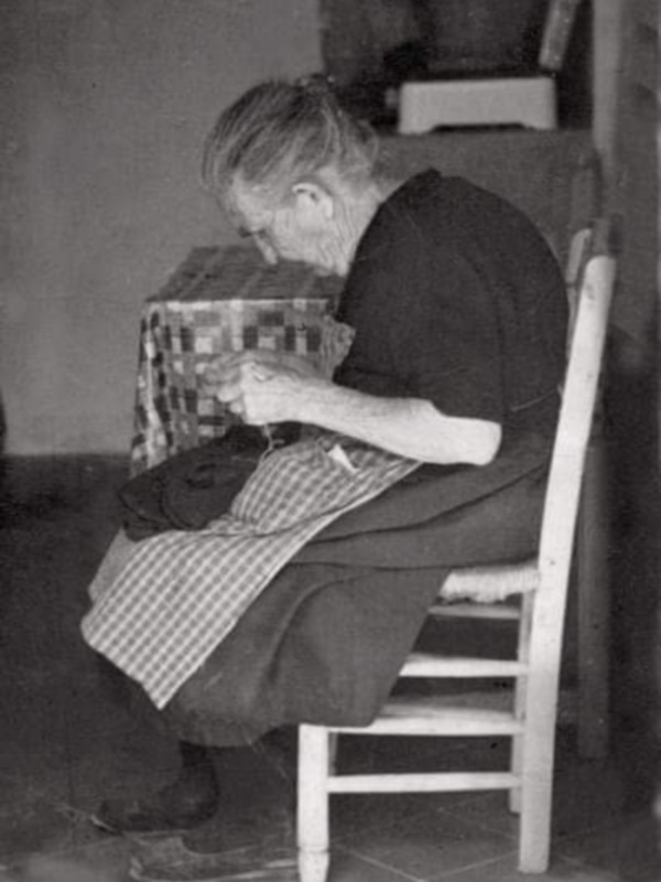 Una anciana sentada en una silla blanca, trabajando con un telar de patchwork.