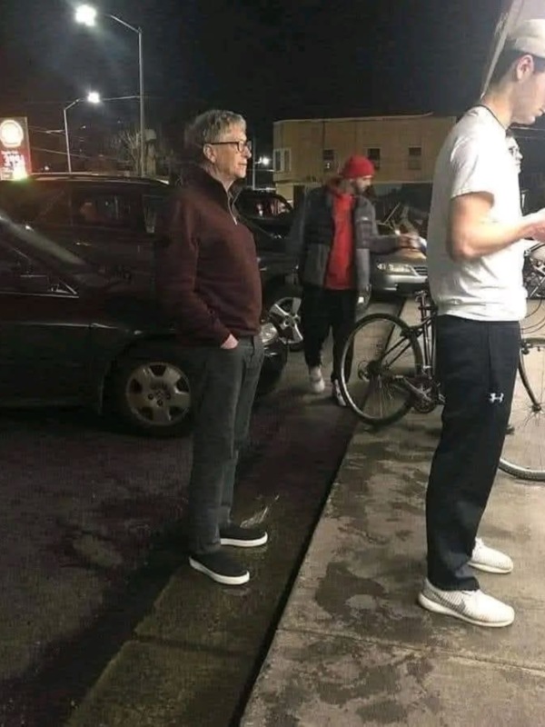 Un hombre en una chaqueta marrón y pantalones va a caminar por la calle de noche, con una bicicleta aparcada al lado.