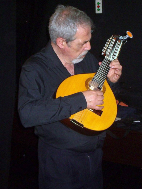 Un hombre con cabello gris y barba blanca toca una guitarra de cuerda doble en un escenario con cortinas negras y luces de fondo.