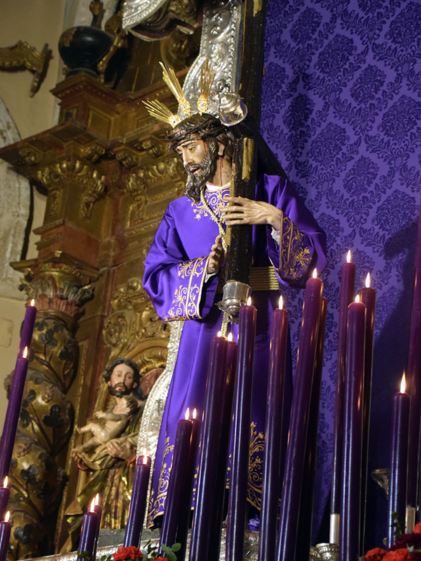 Una estatua religiosa vestida en un manto púrpura con detalles dorados, posiblemente representando a un santo o personaje importante en la religión. La estatua sostiene una cruz y está rodeada por velas encendidas, con un fondo de papel tapiz azul oscuro y detalles dorados en el altar. La escena transmite una atmósfera de reverencia y devoción.