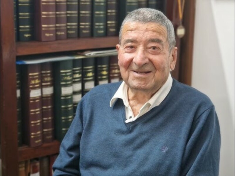 Retrato de un anciano sonriente frente a una estantería de libros.