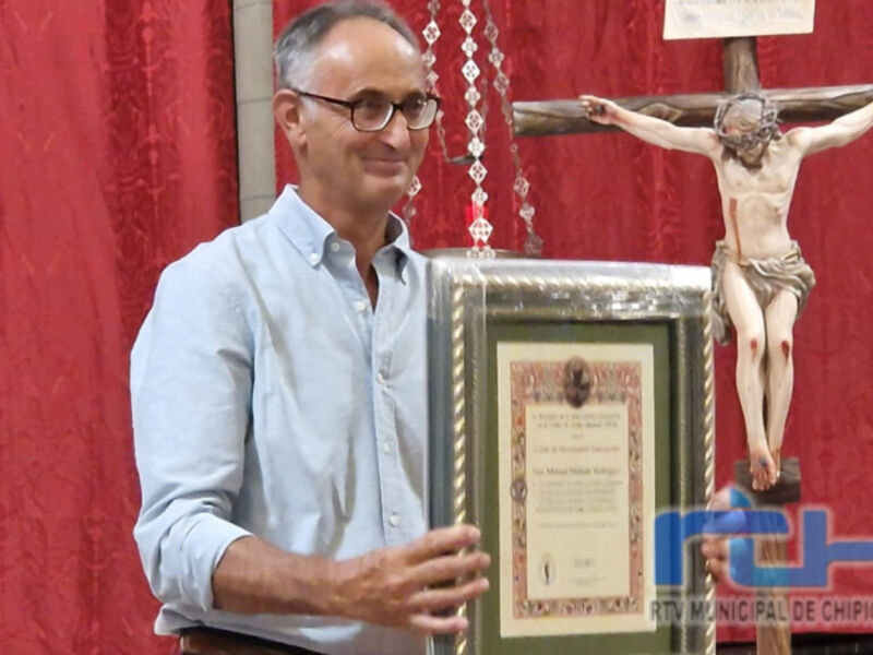Un hombre sostiene un diploma de reconocimiento en una ceremonia religiosa, con un crucifijo en el fondo.