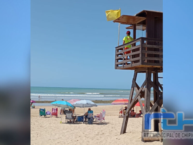Un socorrista en un puesto de vigilancia sobre la playa, con una bandera amarilla al fondo y personas disfrutando del sol en el borde de la arena.