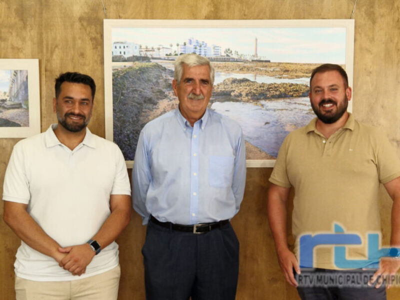 Tres hombres posan frente a una imagen de un puerto, uno con camisa blanca, otro con camisa azul y el tercero con camisa beige. El fondo muestra una vista de un puerto con edificios y agua.