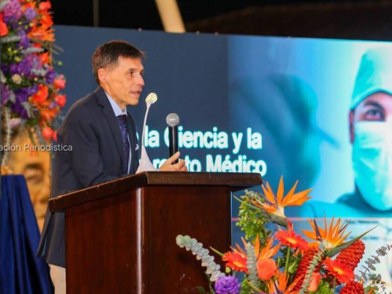 Un hombre habla en un podio con una pantalla de fondo que muestra a un médico. La imagen está relacionada con la innovación periodística y el desarrollo de la ciencia en el campo médico.