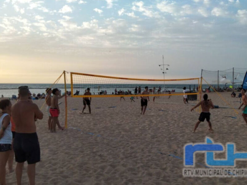 Gente jugando voleibol de playa en la arena con una bandera azul y amarilla en el fondo, durante un evento deportivo organizado por la RTV Municipal de Chipiona.