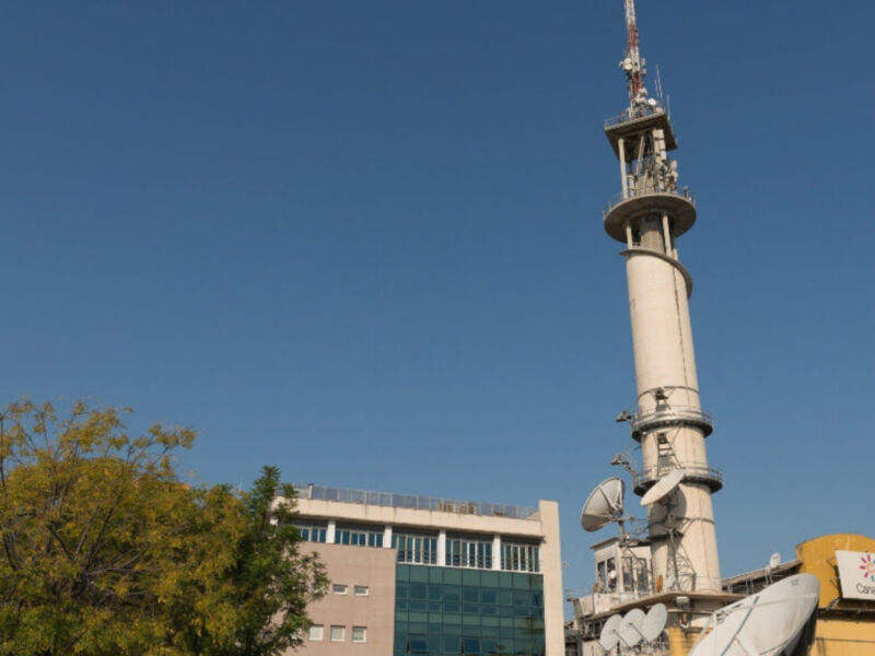 Torre de televisión con antenas y un edificio adyacente bajo un cielo azul claro.