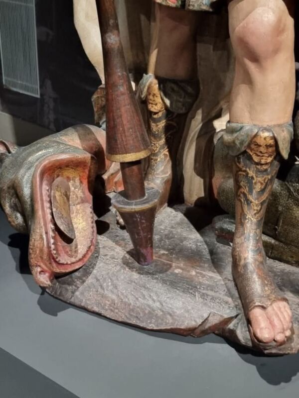 Una escultura de madera tallada con detalles finos y un pie de la figura. La pieza muestra una mano sosteniendo un objeto, posiblemente una vara o herramienta. La madera tiene tonos de rojo y dorado, con detalles en relieve que destacan la textura. La figura parece estar en un entorno interior, posiblemente una exposición o museo.