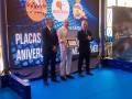 Tres hombres en un escenario azul, uno sostiene una placa de reconocimiento mientras los otros lo observan. El fondo es un gran cartel con logotipos y texto que incluye "Placas Aniversario".