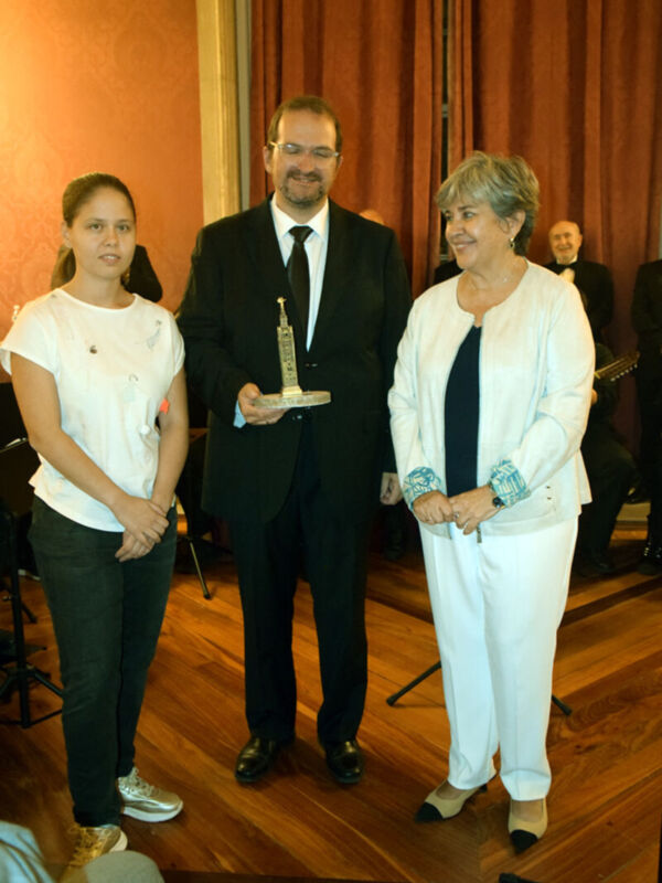 Una imagen de una ceremonia formal donde un hombre sostiene un trofeo mientras una mujer y una niña lo observan. La escena se desarrolla en un ambiente con cortinas rojas y madera, sugiriendo una sala de conferencias o evento.