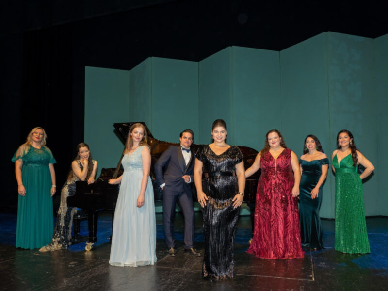 Una escena teatral con siete actores vestidos formalmente, incluyendo un piano en el fondo. La iluminación es dramática con sombras y luces que destacan a los personajes.