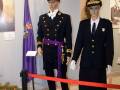 Imágenes de uniformes militares expuestas en una exposición. El hombre lleva un traje negro con detalles dorados y un sombrero de plumas moradas. La mujer está vestida con un uniforme similar, pero en negro y dorado con detalles más suaves. Ambos están frente a una bandera morada.