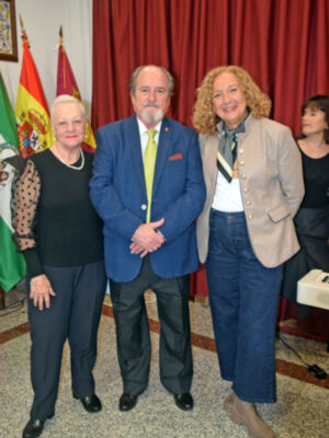 Tres personas posan frente a un fondo de cortinas rojas y banderas. La imagen parece ser de una ceremonia o evento formal, con la presencia de un traje azul y una chaqueta beige.