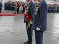 En una sala de conferencias, dos oficiales en uniforme militar se encuentran frente a una fila de soldados. Los oficiales llevan insignias y corbatas, mientras que los soldados están en formación. La escena es formal y ceremonial.