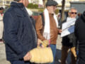 Una calle con gente caminando, algunos con gorras y chaquetas. Un hombre tiene una botella amarilla y un papel en la mano. Fondo de edificios, árboles y cielo azul claro.